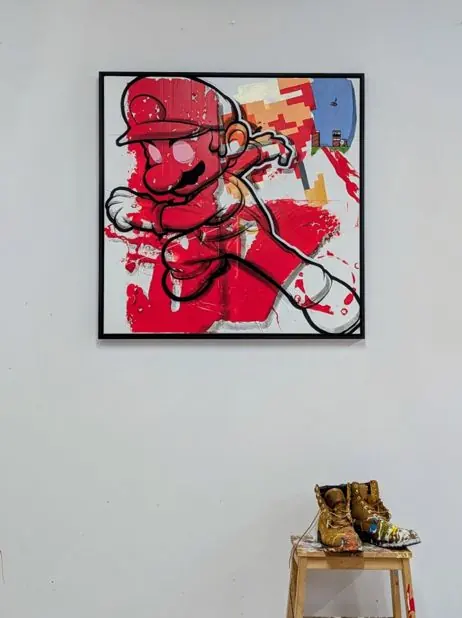 Jumpman by Remco, a square mixed media painting in reds with an 8-bit game scene, hung on a white studio wall above paint-spattered work boots