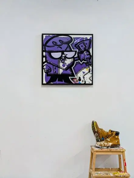 The Boy Genius by Remco, a square purple mixed media painting hung on a white studio wall beside paint-spattered work boots