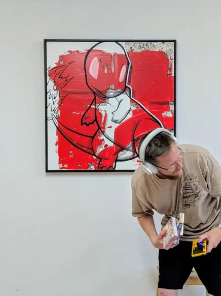 The Red Starter by Remco, a square red mixed media painting on a white studio wall, with the artist in headphones standing beside it holding a handheld games console